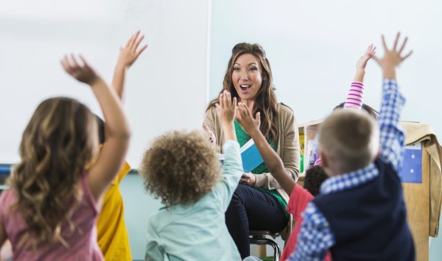 A female, Asian preschool or kindergarten teacher in the classroom, sitting in front of a group of multi-ethnic children, reading a book.  The boys and girls are raising their hands to answer a question.  There is a large, blank whiteboard in the background for copy.